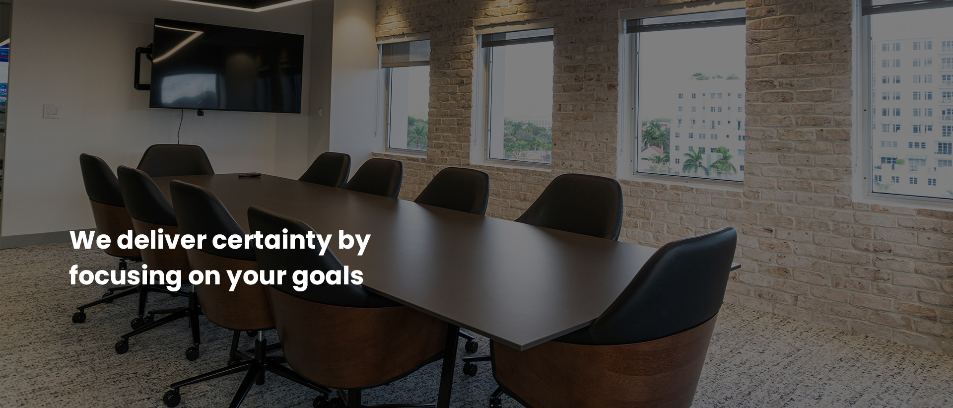 Conference room with a long dark table and leather chairs beside an exposed brick wall and city windows; the image carries the caption “We deliver certainty by focusing on your goals”