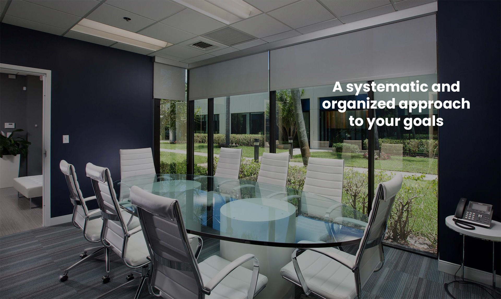 Glass-walled conference room with a white table, white chairs and roller shades looking onto a garden courtyard; the image carries the caption “A systematic and organized approach to your goals”