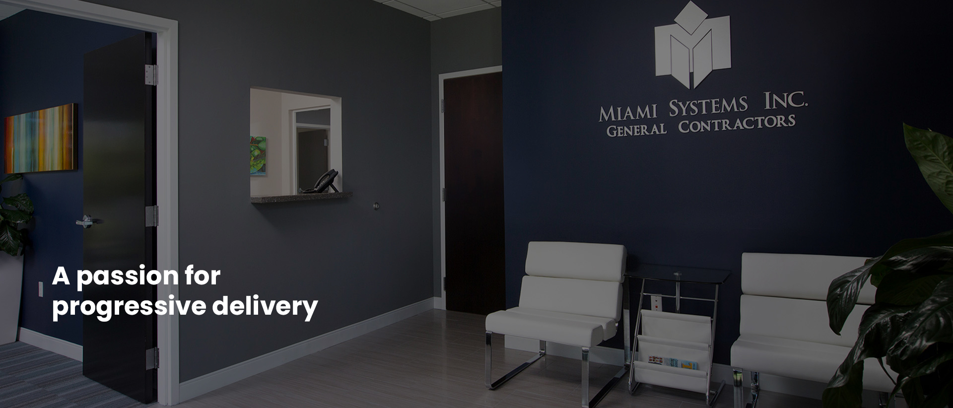Office reception with a dark feature wall bearing the Miami Systems Inc. General Contractors sign, a seating area and a doorway to a private office; the image carries the caption “A passion for progressive delivery”